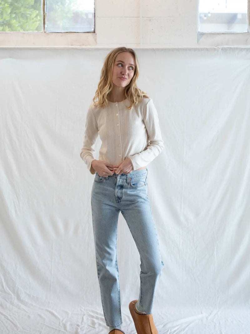 Woman wearing a light-colored shirt and blue jeans standing against a white wall.