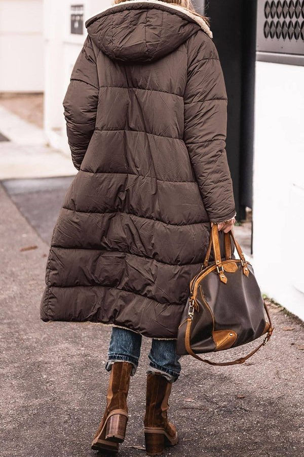 Person wearing a long dark brown puffer coat and holding a brown handbag on a street.