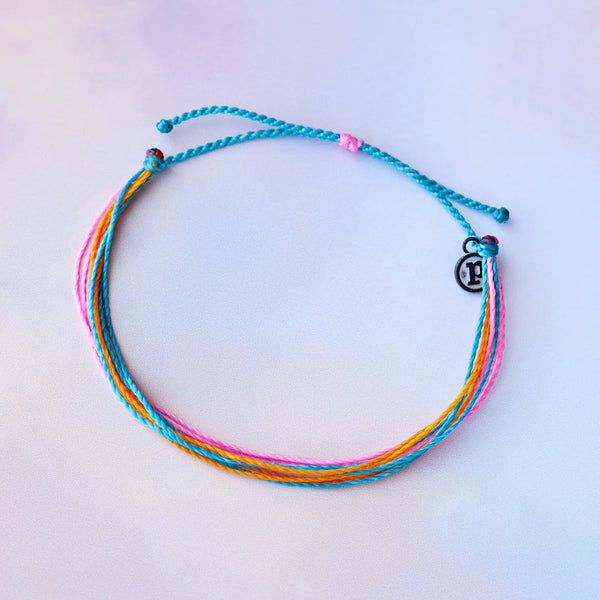 shows a tie dye backdrop under anklet made of teal, pink, and orange wax coated strings. features a metal disc charm with a "p" inside