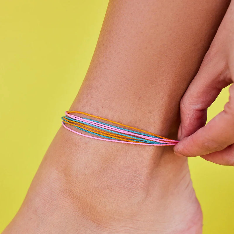 Shows a yellow backdrop with an ankle wearing an anklet made of teal, pink, and orange wax coated strings