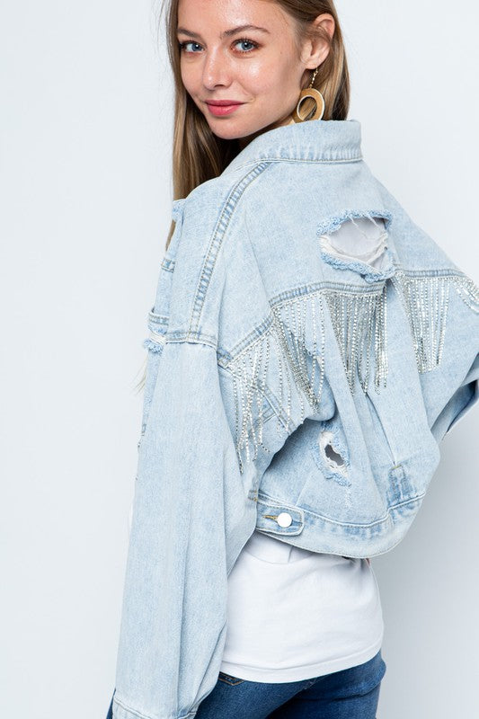 Model turn to look back at camera, is wearing rhinestone fringe distressed denim jacket which falls above the waist- sleeves are long and past her hands