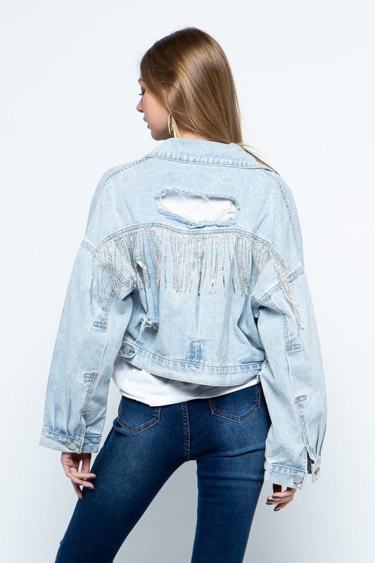 model is turned away showcasing back of jacket where you can see a fringe across back of jacket as well as a distressed hole in the light wash denim, she is wearing a white tshirt and dark skinny  jeans