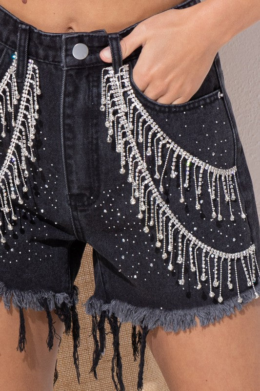 close up view of black denim shorts with allover sparkle detail and rhinestone chains hanging from sides, frayed hemline and functional pockets