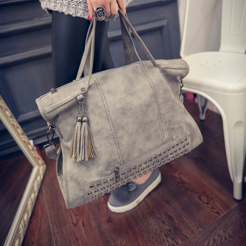 Front view of handbag. Shows the studded detail. Also shows the two-tassel detail, the short handles & the zipper closure.   