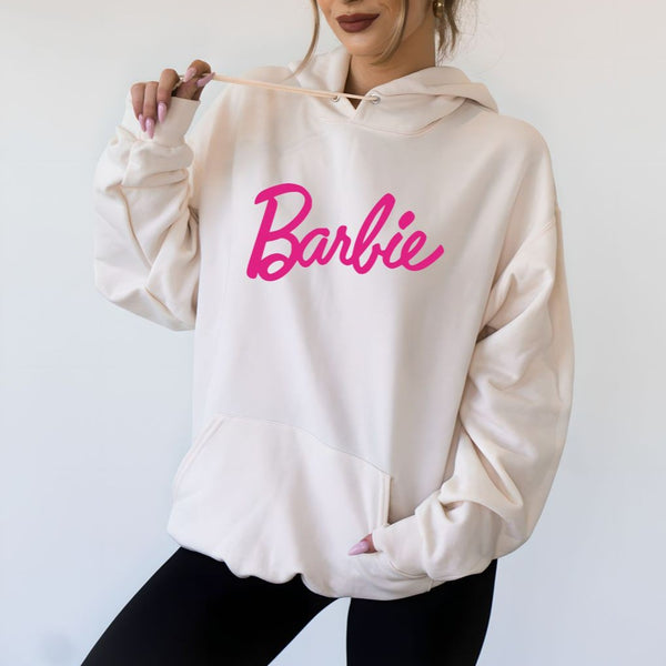 front view of model in cream hoodie. shows the front pouch pocket, drawstring at neckline, dropped shoulders and cursive "barbie" in pink across the front.