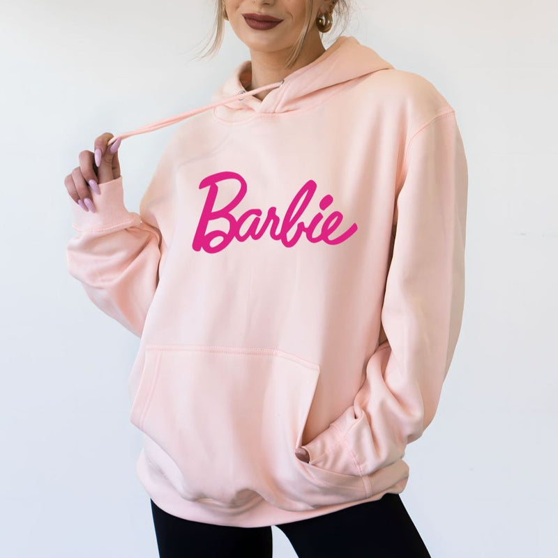 front view of model in light pink hoodie. shows the front pouch pocket, drawstring at neckline, dropped shoulders and cursive "barbie" in pink across the front.
