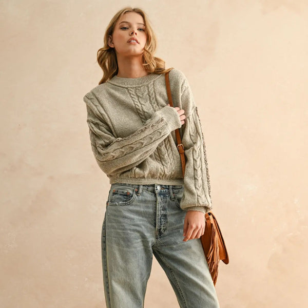 Woman wearing a cable knit sweater and jeans holding a brown leather bag against a beige background