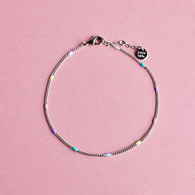 Shows a pink backdrop with a silver chain anklet lined with light blue, teal, purple, pink, and yellow beads. features a lobster closure, 2 extensions, and a round charm etched with "pura vida" on the end.