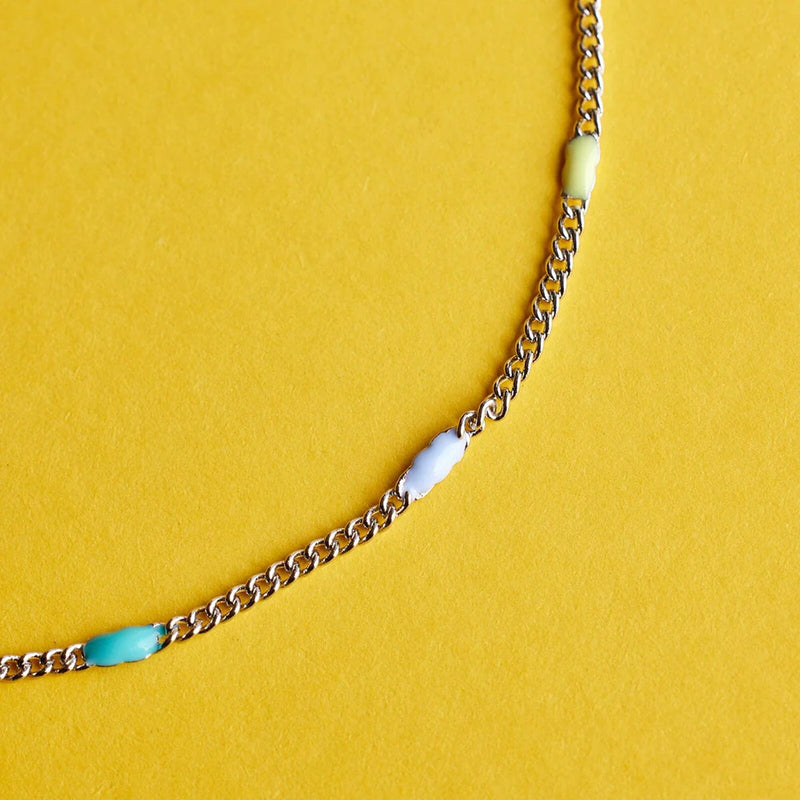 shows a yellow backdrop under a close up of a silver chain anklet lined with yellow, light blue, and teal enamel beads 