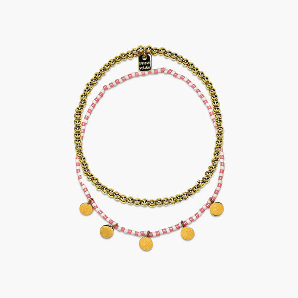 Shows both of the bracelets in the gold set laying on a flat surface. Stretch pull on design. One bracelet is solid gold beads and the other is a pink and white beaded bracelet with flat circular beads hanging off.