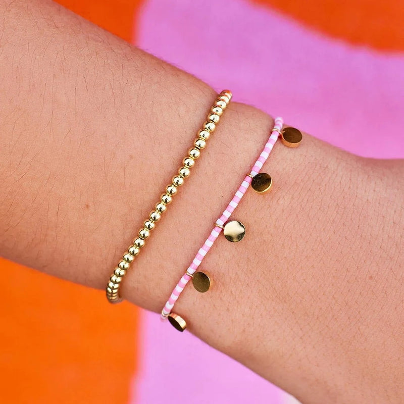 Model is wearing the gold 2 bracelet set. One bracelet is solid gold beads and the other is a pink and white beaded bracelet adorned with flat circle charms hanging off. 