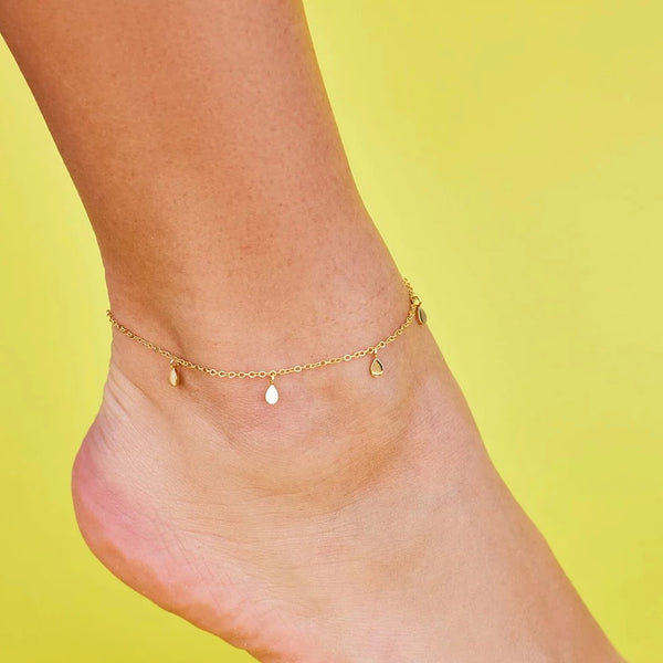 Gold chain anklet is shown on models ankle. Features thin gold chain with small, spaced out gold teardrop charms hanging from it.