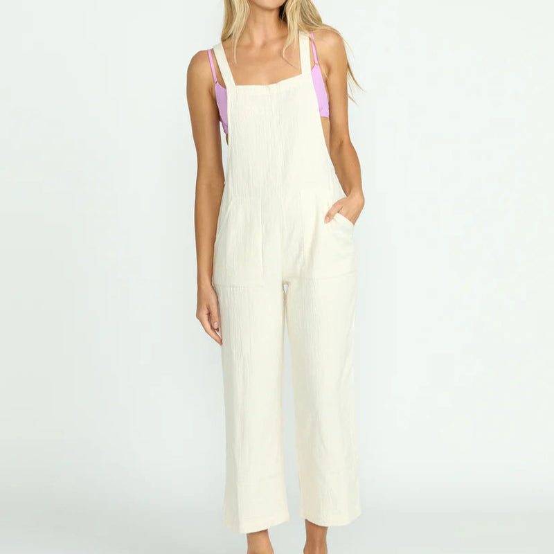 front view of the model wearing the pacific time jumpsuit. shows the straight neckline. also shows patch pockets, the straps and the white color. 