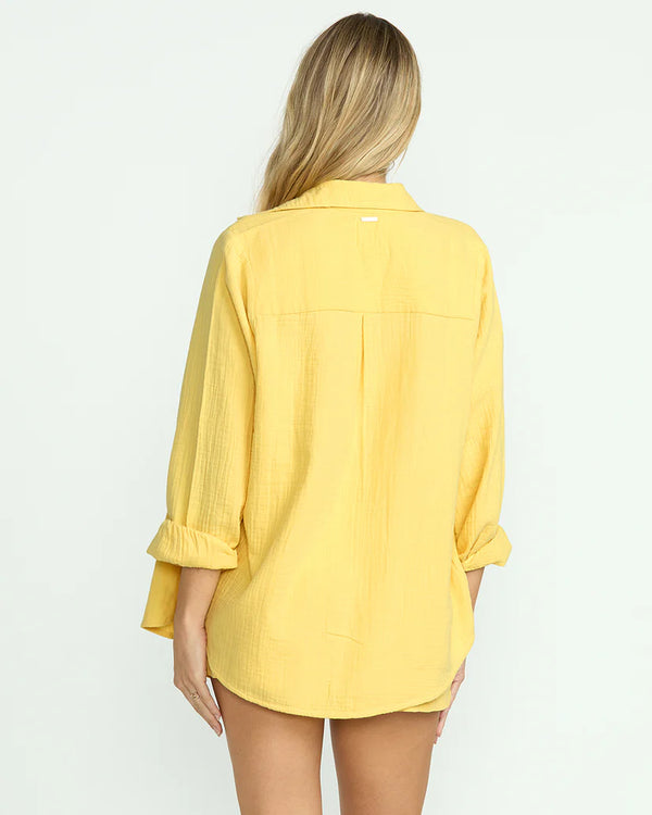 back view of the model wearing the sweet swell long sleeve shirt. shows the metal plate. also shows the collar, the oversized fit and the yellow color. 