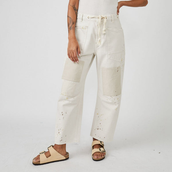 front view of model in baggy pants. shows tie waist with gromet holes, large front pockets, patches on knees and paint splatter. the legs are wide and hit model at ankle.