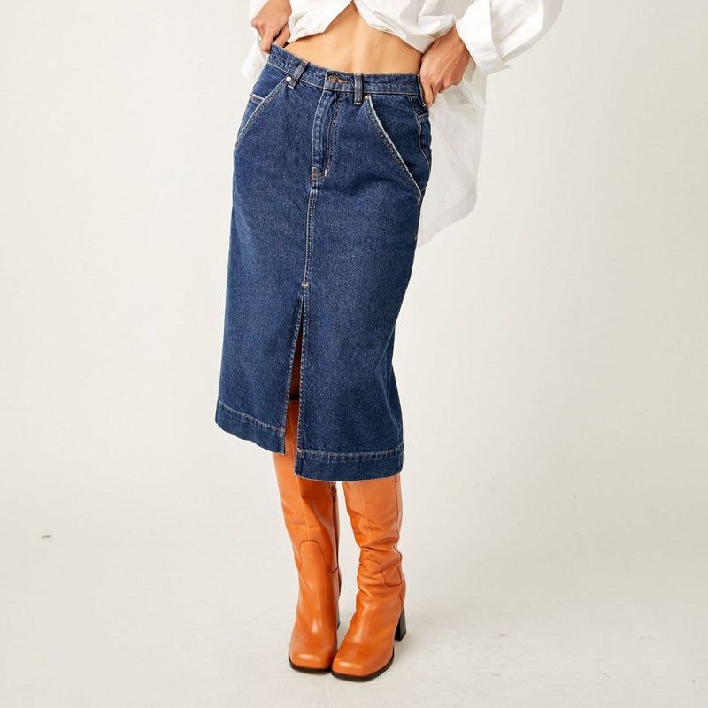 front view of model in skirt. shows rigid denim fabrication, mid waist, side pockets, a front slit hemline and midi length.