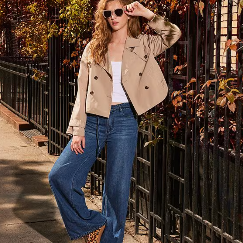 model stands with one leg propped and leaning against a fence, jeans are shown to be wide and flowing from the knee down