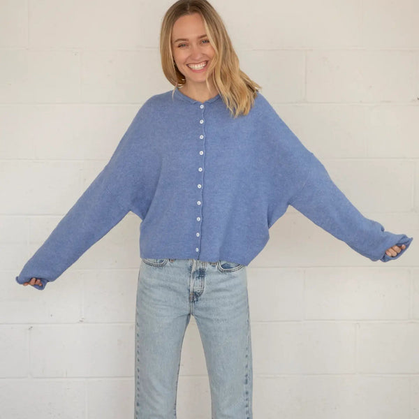 Woman wearing a blue cardigan and jeans against a white brick wall.