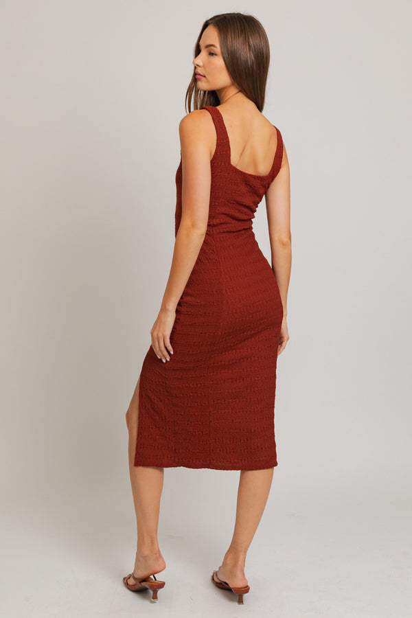 Back view of model wearing dress. Shows the textured detail throughout the dress. Also shows the back square neckline, you can see the front slit from this angle, the midi length and the red/brick color of the dress.