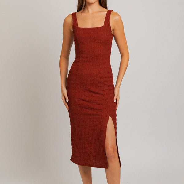 Front view of model wearing dress. Shows the tank style of the dress. Also shows the square neckline, the front slit, the textured detail throughout them dress, the midi length and the color of the dress is this red/brick color. 