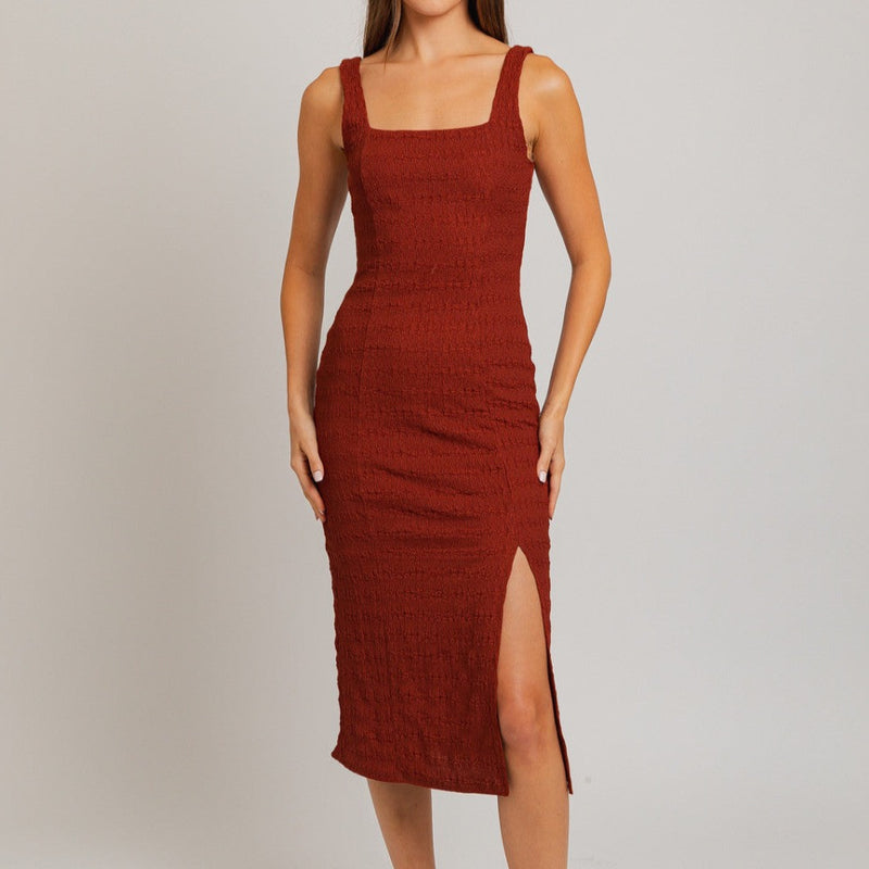 Front view of model wearing dress. Shows the tank style of the dress. Also shows the square neckline, the front slit, the textured detail throughout them dress, the midi length and the color of the dress is this red/brick color. 