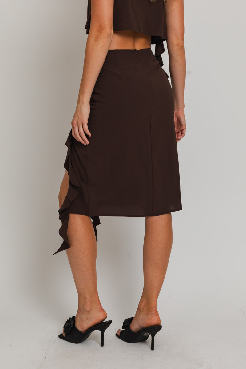 back view of model wearing skirt. Shows the low rise of the skirt. Also shows the front slit with the ruffle from the front of the skirt, the knee length of the skirt and the color of the skirt is dark brown and the the back zipper closure. 