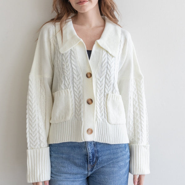 front view of model wearing the callow sweater in ivory. shows the collar. also shows the two. front patch pockets, the folded cuffs, ribbed detail & the wood buttons. 