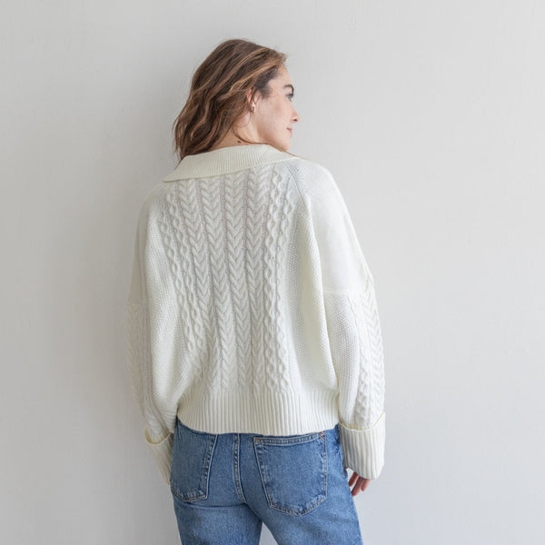 back view of the model wearing the clue sweater in ivory. shows the collar. also shows the ribbed detail, the cable knit detail through the back and the folded cuffs/