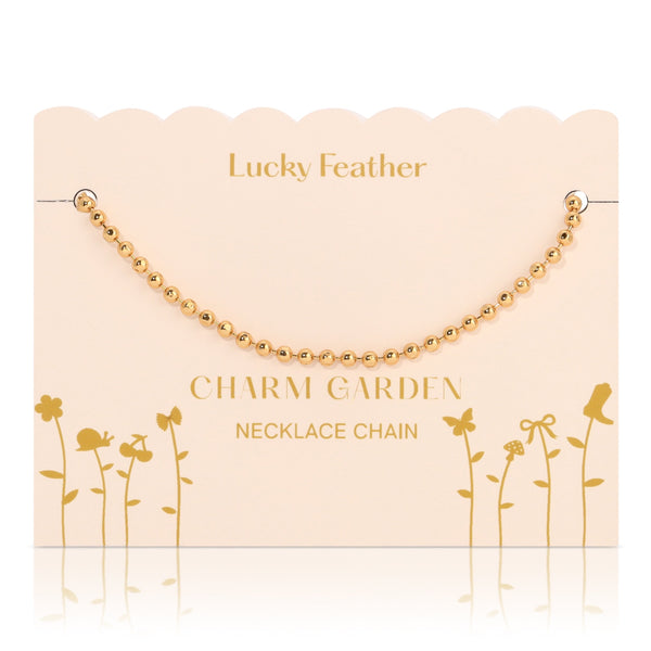 front view of gold necklace chain on the light pink packaging with gold wording LUCKY FEATHER CHAIN GARDEN NECKLACE CHAIN. In the middle Is the gold necklace chain. The chain is little round gold balls. 