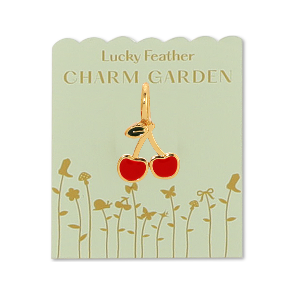 Front view of charm on packaging. Shows the cherry charm, also shows the light green packaging with gold flowers and gold writing lucky feather charm garden. Charm is a little bit smaller than a dime.