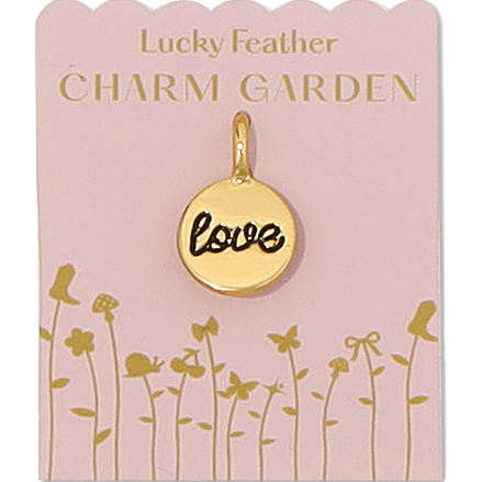 Front view of LOVE gold charm on pink packaging with gold flower detail and gold wording lucky feather charm garden. Shows the gold love charm, the word love is in cursive and black writing on front of charm. Charm is a little bit smaller than a dime. 