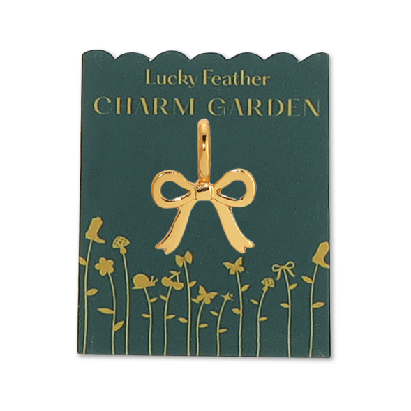 Front view of bow charm on packaging. Shows the gold bow charm. Also shows the green packaging with gold flowers and the wording saying LUCKY FEATHER CHARM GARDEN.