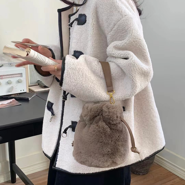 side view of model wearing the daphne fur bucket crossbody in taupe. shows one side of the pull closure, the shoulder strap, the gold hardware, and the faux fur body. 