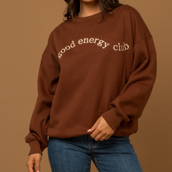 front view of the Helen good energy club sweatshirt in brown. shows the crew neckline. also shows the drop shoulders, the wording good energy club on the front in cream and the oversized fit. 