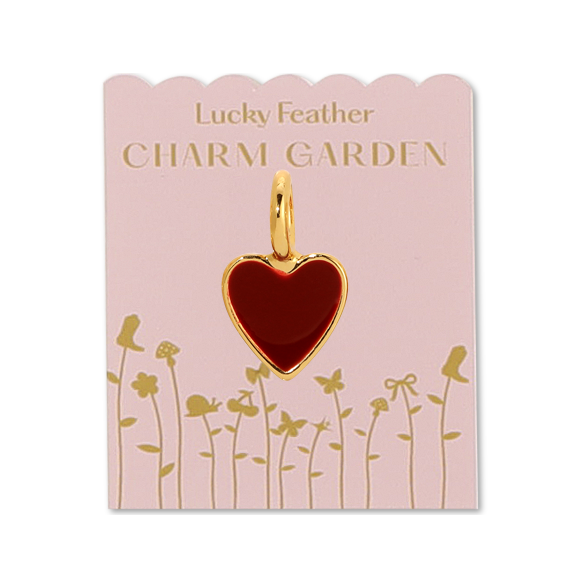 Front view of gold heart charm on the pink packaging with gold wording LUCKY FEATHER CHARM GARDEN and gold flowers on the bottom of the packaging. Shows the gold heart charm that is red in the middle of the packaging. 