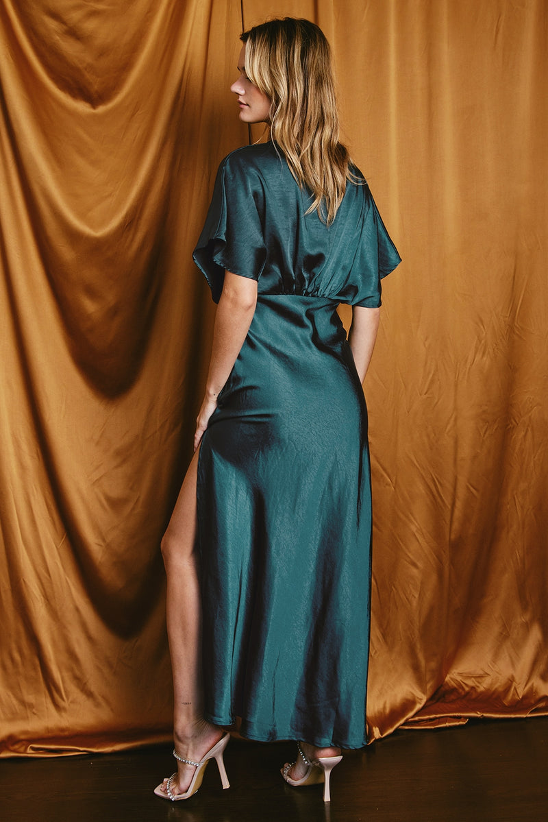 back view of the model wearing the mallory maxi dress. shows the side slit. also shows the satin look, the blouson top and the short sleeves. 