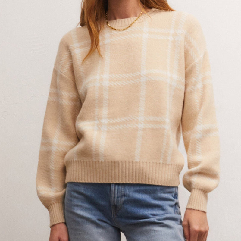 Front view of model wearing sweater. Shows the ribbed detail on the neckline, cuffs and hem. Also shows the plaid knit pattern and the waist length of the sweater. The color of the sweater is an almond color with plaid being white. 