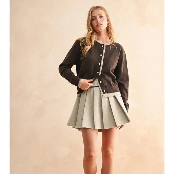 Woman wearing a brown cardigan, black top, gray pleated skirt, and black boots against a beige background