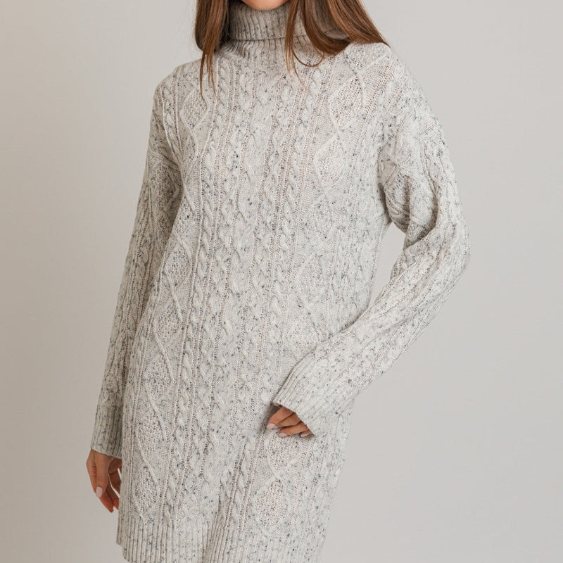 Front view of model wearing dress. Shows the cable knit detail. Also shows the turtleneck, the ribbed detail on the turtleneck, cuff and bottom hem. Shows the speckling in the fabric. 