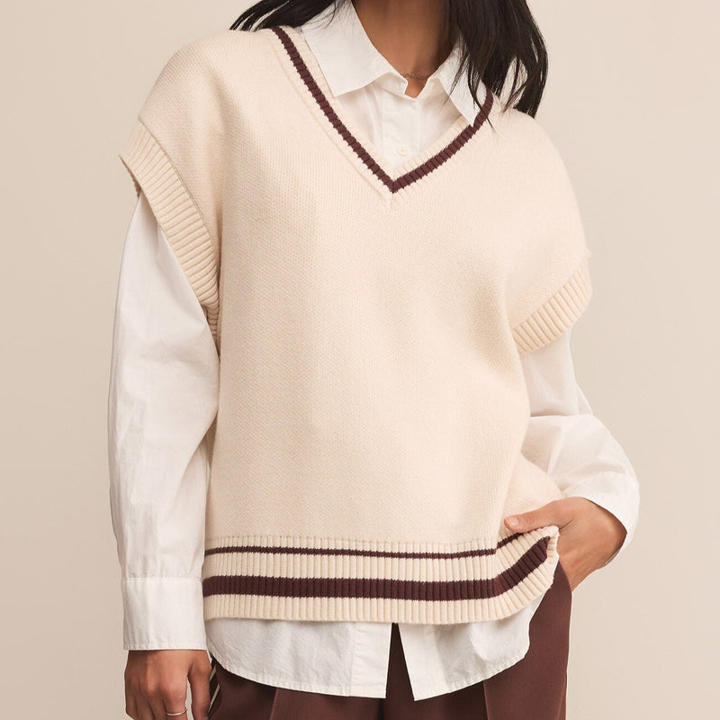 Woman wearing a cream sweater with a V-neck and brown trim over a white shirt, standing against a beige background.