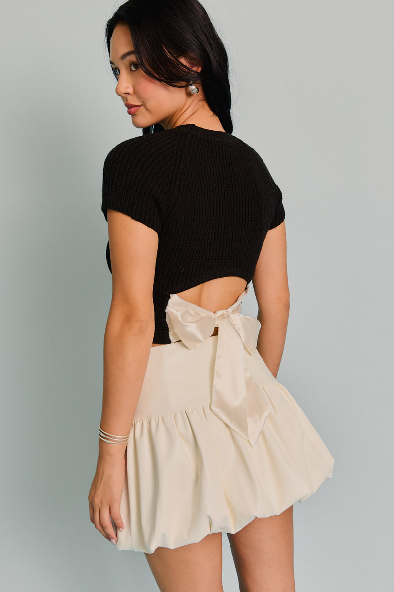 back view of the model wearing the becky knit top. shows the back tie detail. also shows the tie is a cream color and the shirt is all black, shows the short sleeves and the cropped length. 