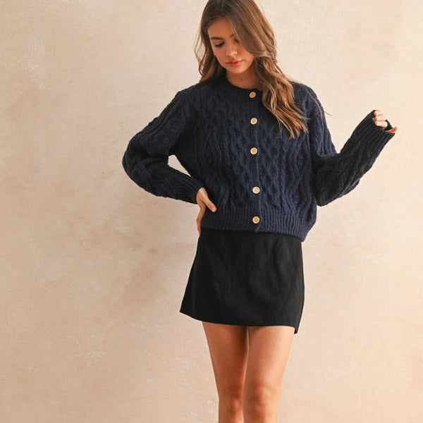 Woman wearing a navy blue cable knit cardigan with gold buttons, black skirt, and black shoes against a beige wall.