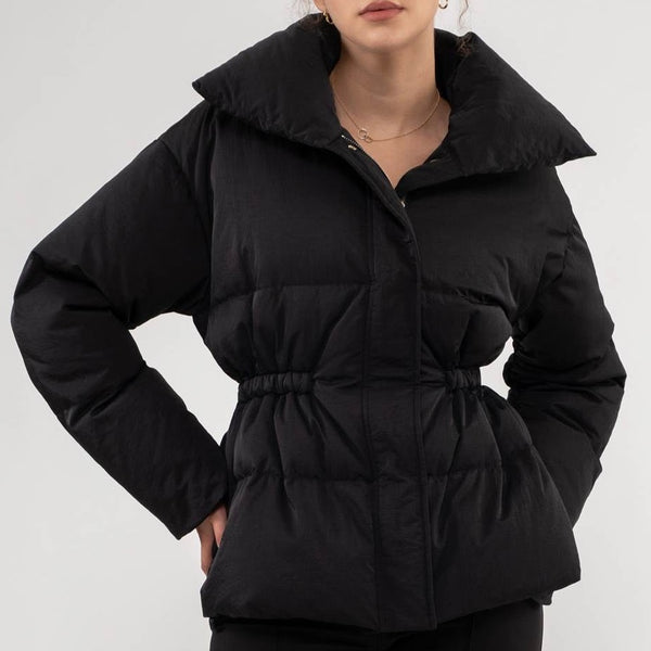 front view of the model  wearing the Kelly puffer jacket in black. shows the exaggerated collar. also shows the elastic waist, the zipper/button closure and the puffer texture.