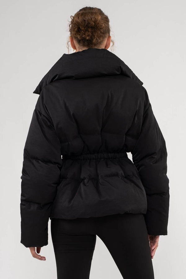 back view of model wearing the Kelly puffer coat in black. shows the elastic waistband. also shows the exaggerated collar and the puffer texture.