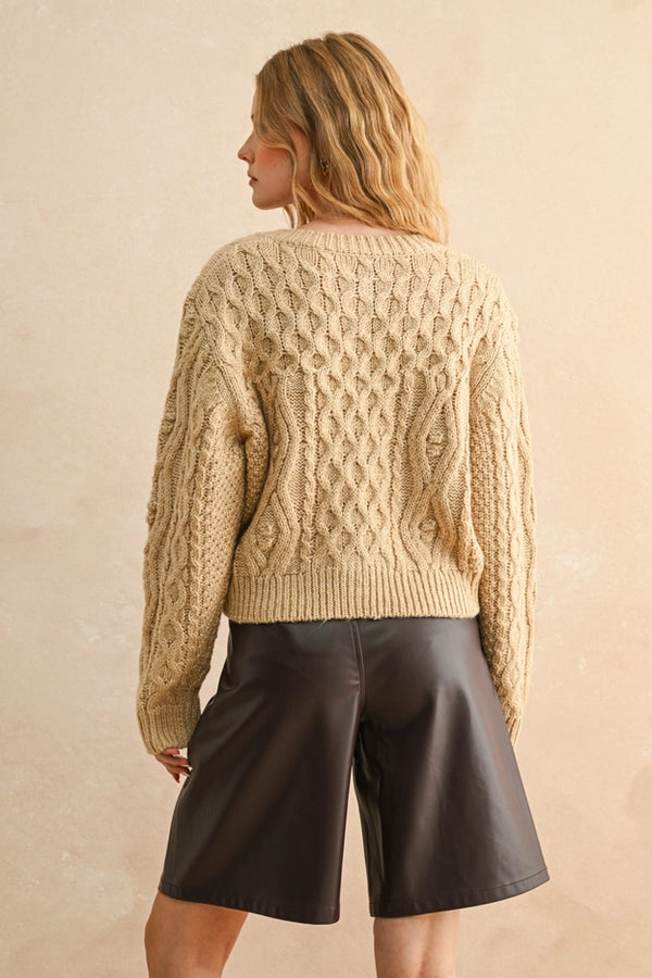 Woman wearing a beige cable knit sweater and dark shorts against a beige background
