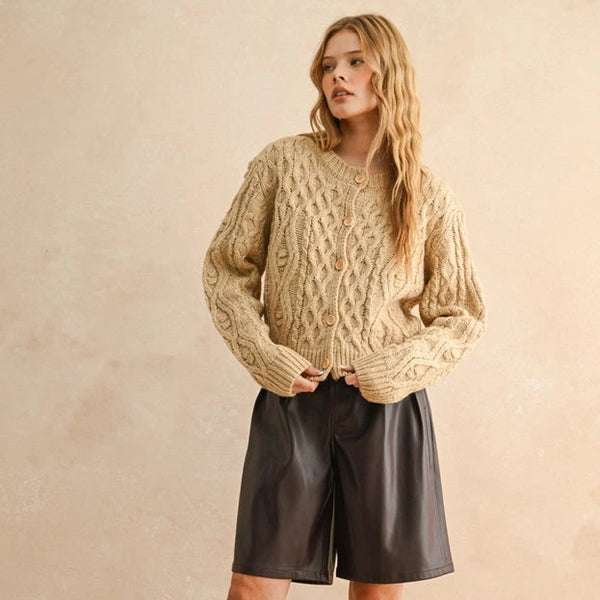 Woman wearing a beige cable knit sweater, black leather shorts, and black boots against a beige background