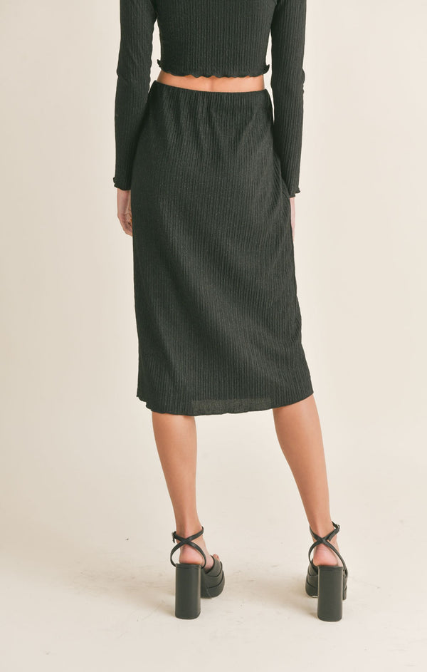 back view of model wearing the Nelly midi skirt. shows the mid length. also shows the ribbed detail, the high waist and the elastic waistband. 