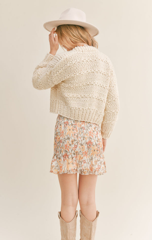 Back view of the model wearing the cardigan. Shows that cardigan is cropped  and that the cardigan bans at the cuffs on the sleeves. 
