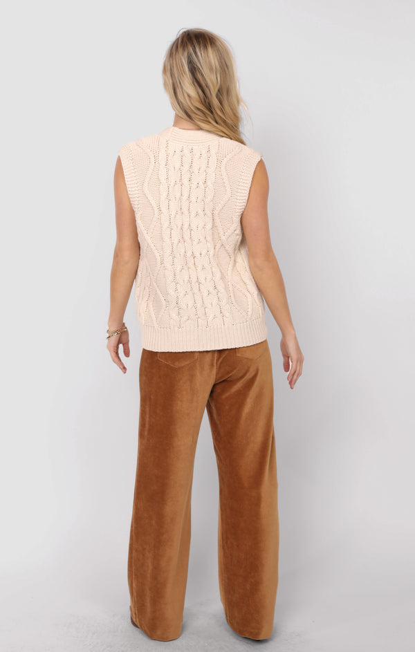 Person wearing a cream sleeveless sweater and brown pants on a white background