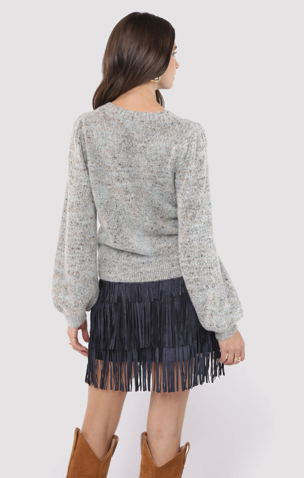 Woman wearing a gray sweater and black fringe skirt with brown boots on a white background